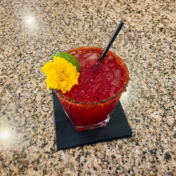 Red cocktail with a chili-lime rim, garnished with a lime wedge and yellow flower on a granite counter.