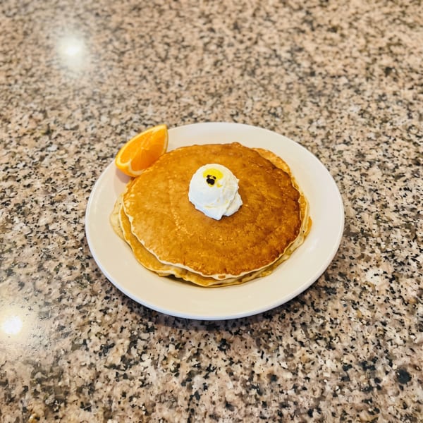 Golden pancakes topped with whipped cream, a pat of butter, and an orange slice on a white plate.
