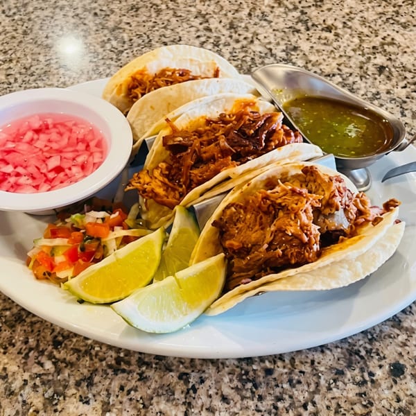 A plate of four tacos with shredded meat, lime wedges, salsa, and pickled onions.