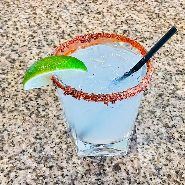 A light blue cocktail with a chili-lime rim, lime wedge, and black straw on a granite surface.