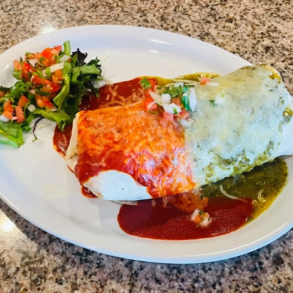 A large burrito covered in red and green sauce, with a side of salad, on a white plate.