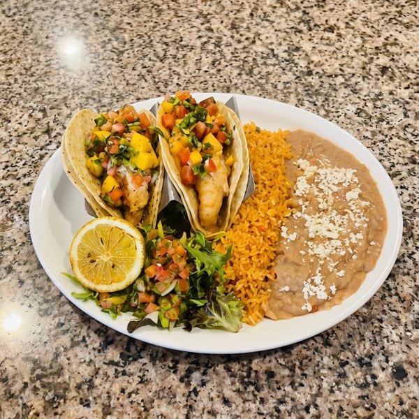 Two shrimp tacos with mango salsa, rice, refried beans, and a lemon slice on a white plate.